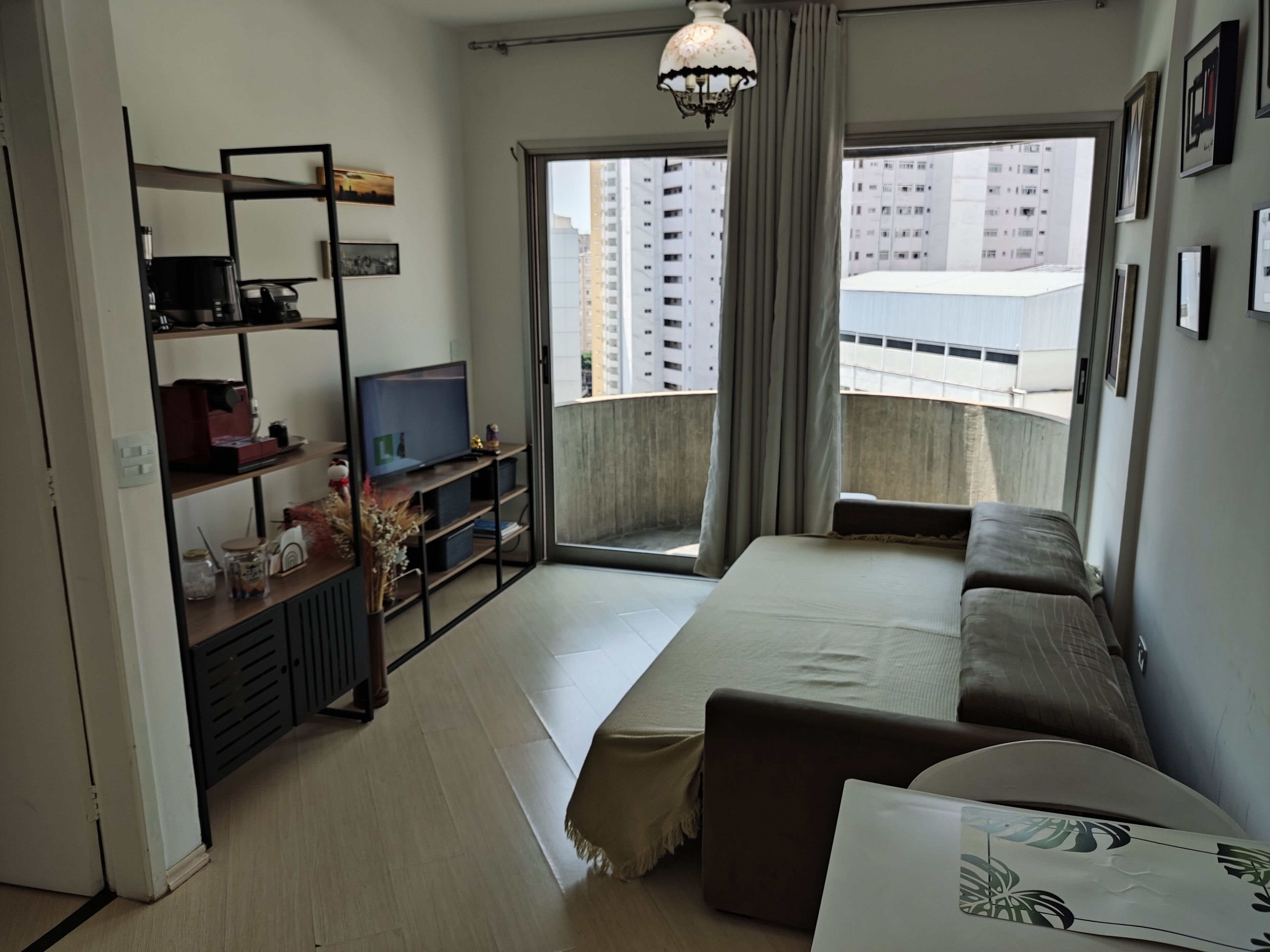 A modern living room with a sofa, a TV on a black stand, a bookshelf, and a balcony with a view of high-rise buildings.