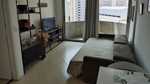 A modern living room with a sofa, a TV on a black stand, a bookshelf, and a balcony with a view of high-rise buildings. - APARTAMENTO ACONCHEGANTE LIBERDADE SP (São Paulo)