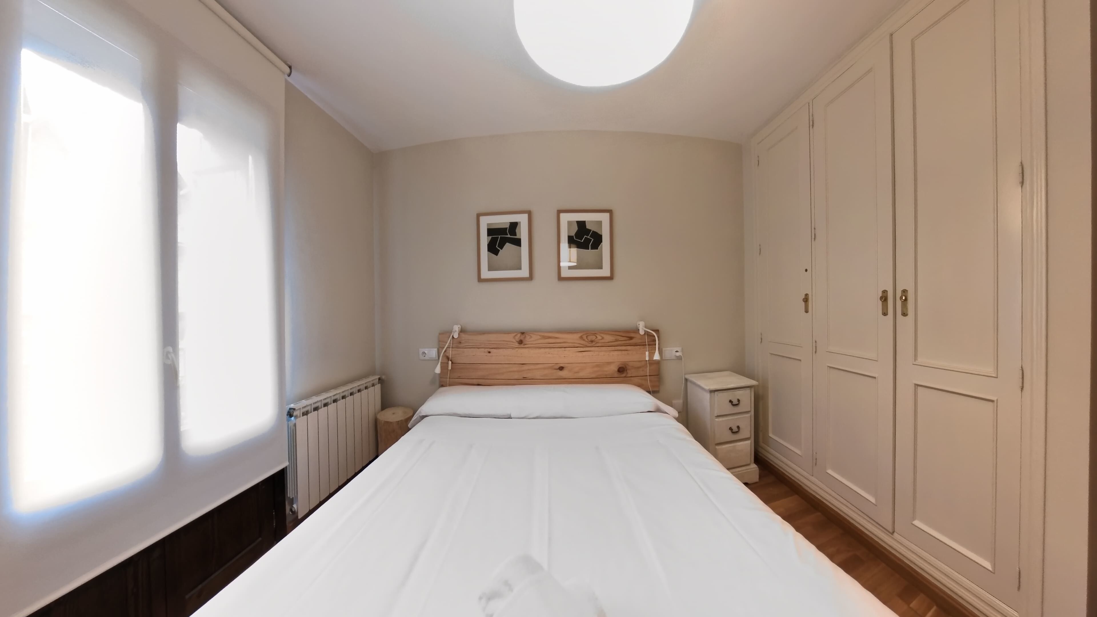 A bedroom with a wooden headboard, a white bedspread, a bedside table with drawers, a radiator, and a wardrobe.