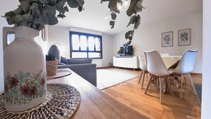 Ein modernes Wohnzimmer mit einer Couch, einem Essbereich mit Stühlen, einer großen Vase mit Blumen und Blick durch ein Fenster nach draußen.