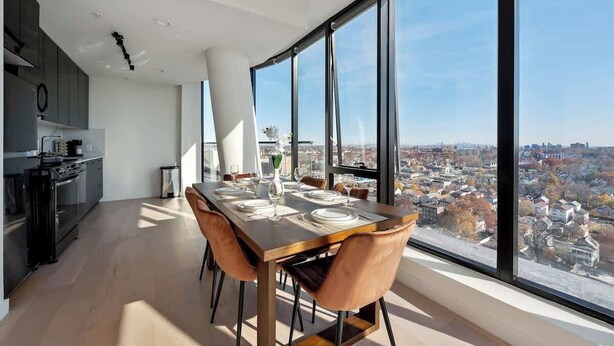 Un espace de repas moderne avec une grande table dressée pour quatre personnes, une vue sur la ville à travers de grandes fenêtres et une cuisine dotée de meubles sombres.