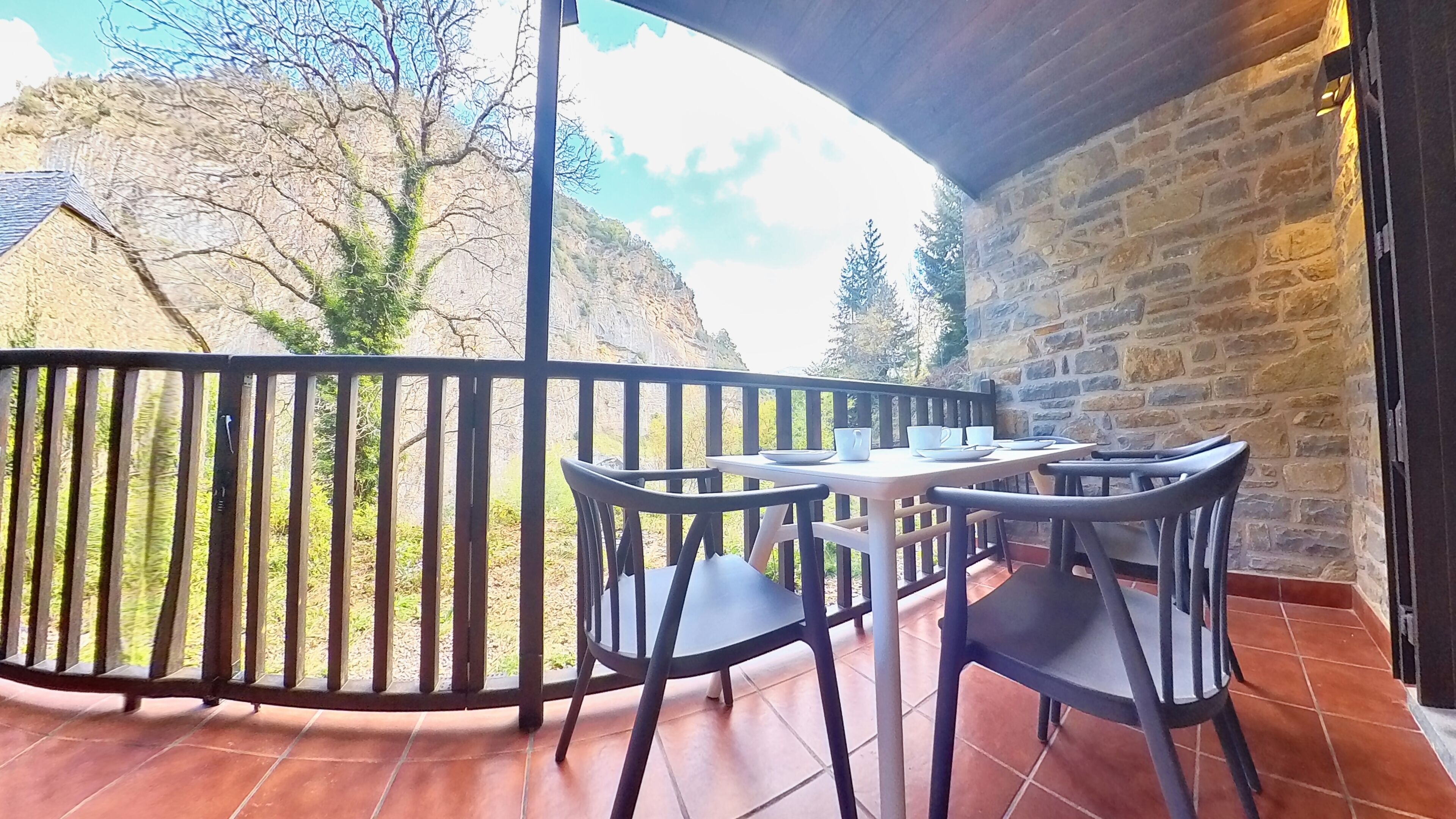 A balcony with a stone wall, a table with chairs, and a view of a mountain and trees.