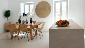 Un coin repas avec une table en bois et des chaises en osier, une grande tapisserie murale tissée et un panier de fruits posé sur un comptoir.
