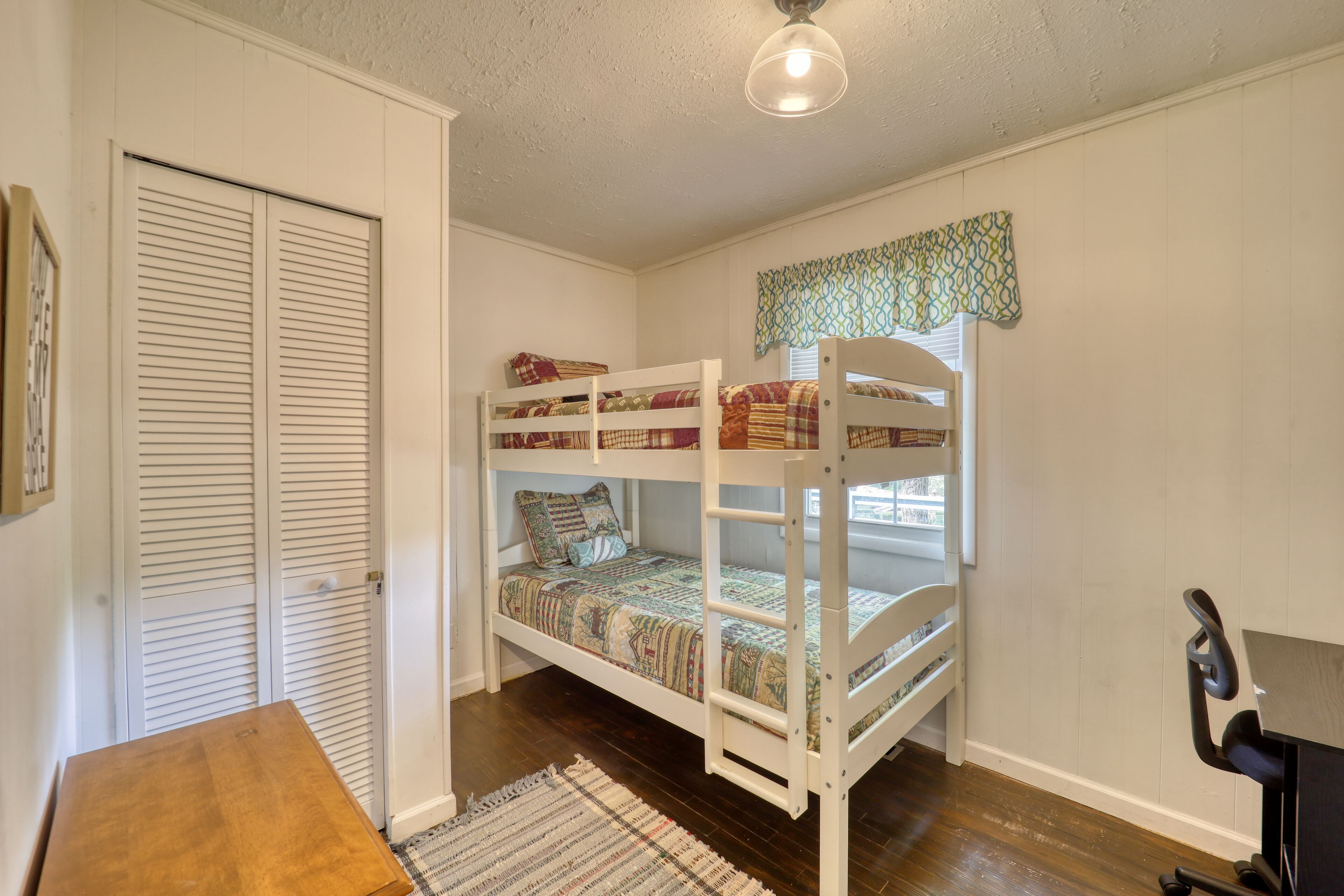 Une chambre avec un lit superposé, un bureau et une chaise, un tapis à motifs et une fenêtre agrémentée d’une lambrequin à motifs.