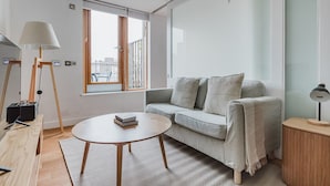Un salon moderne avec un canapé gris, une table basse ronde, un lampadaire et une fenêtre donnant sur des immeubles.