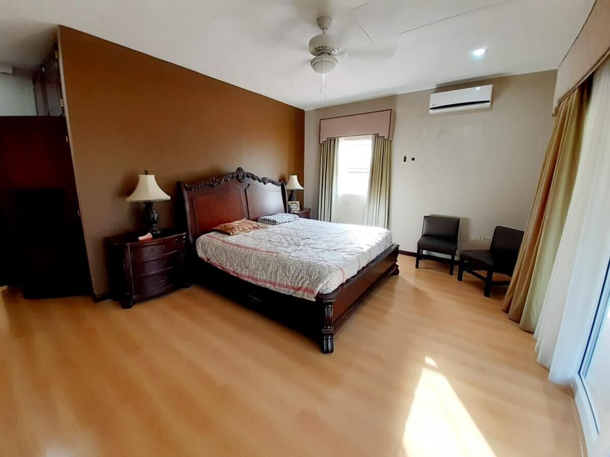 Un dormitorio con una cama grande, dos mesitas de noche, un ventilador de techo, un aire acondicionado y una ventana con cortinas.