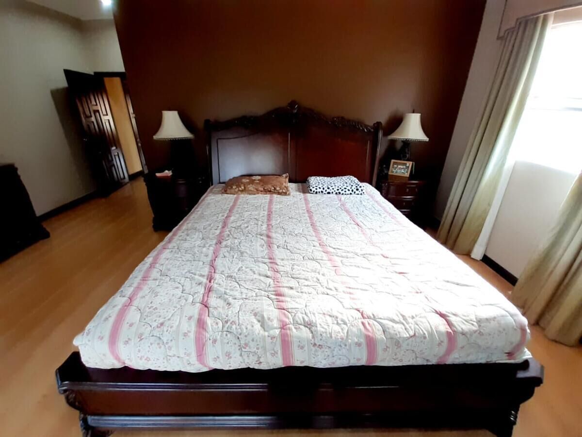Una cama bien tendida con una colcha floral y dos almohadas, un cabecero de madera, dos mesitas de noche con lámparas y vistas a una ventana con cortinas.