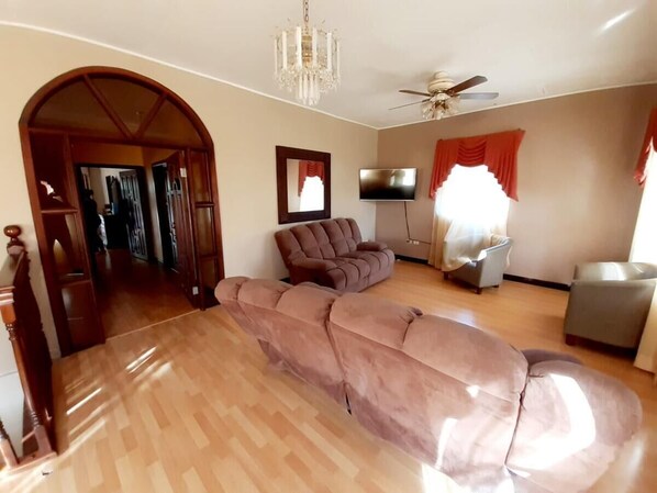A spacious living room with a wooden floor, a brown sofa, a grey armchair, a ceiling fan, and a TV.