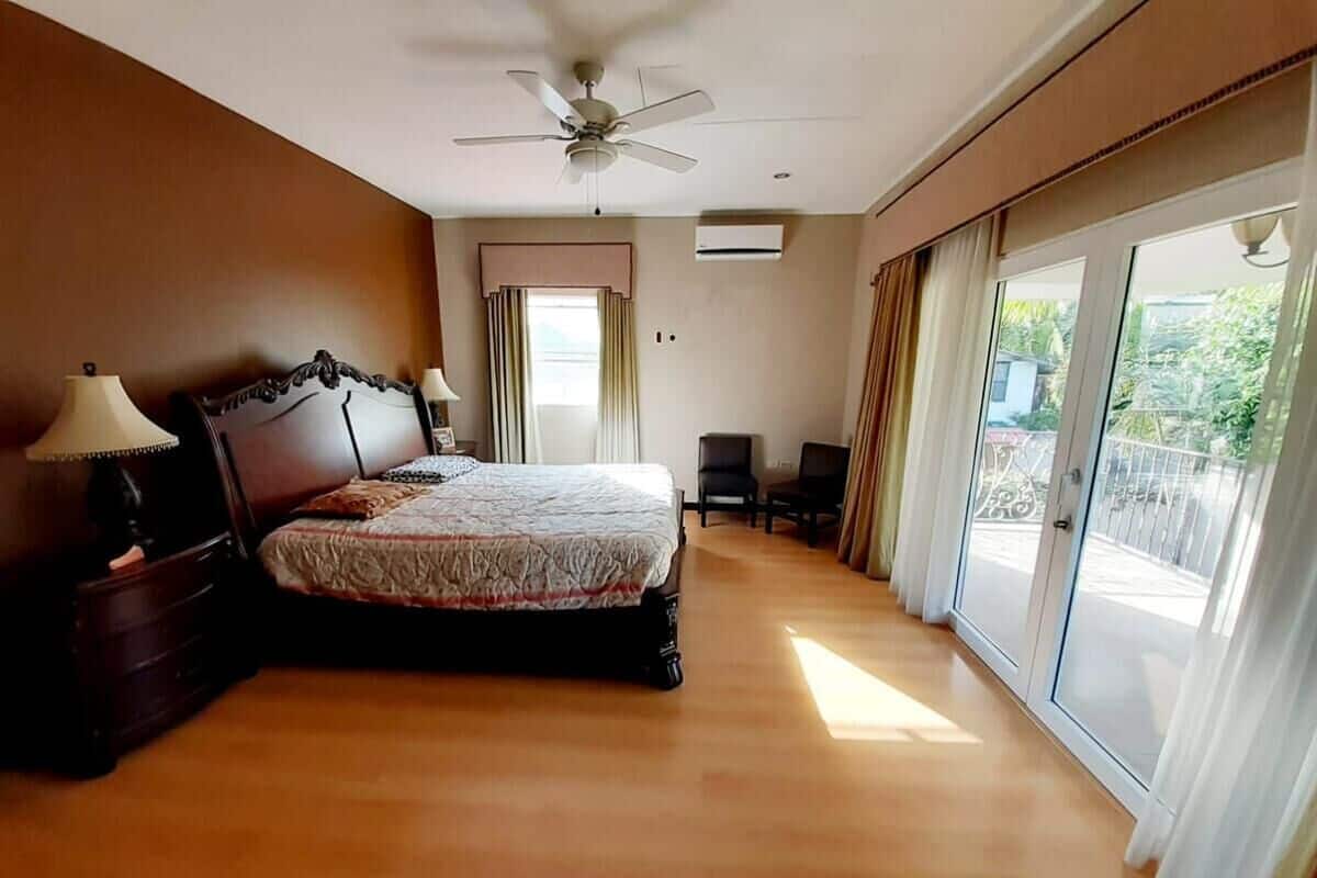 A bedroom with a bed, a nightstand, a lamp, a ceiling fan, and a view of the outside through large windows.