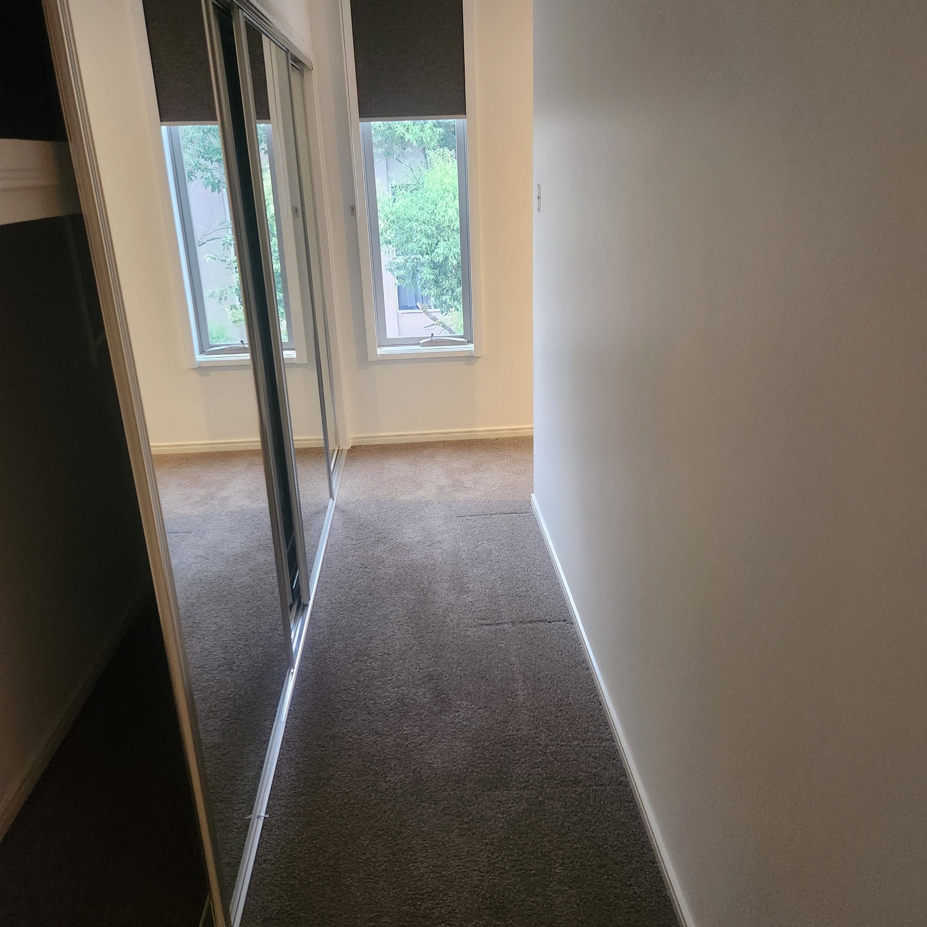 A narrow hallway with carpeted floors and white walls, leading to a window with a view of greenery outside.