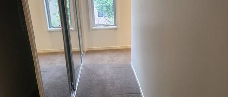 A narrow hallway with carpeted floors and white walls, leading to a window with a view of greenery outside.
