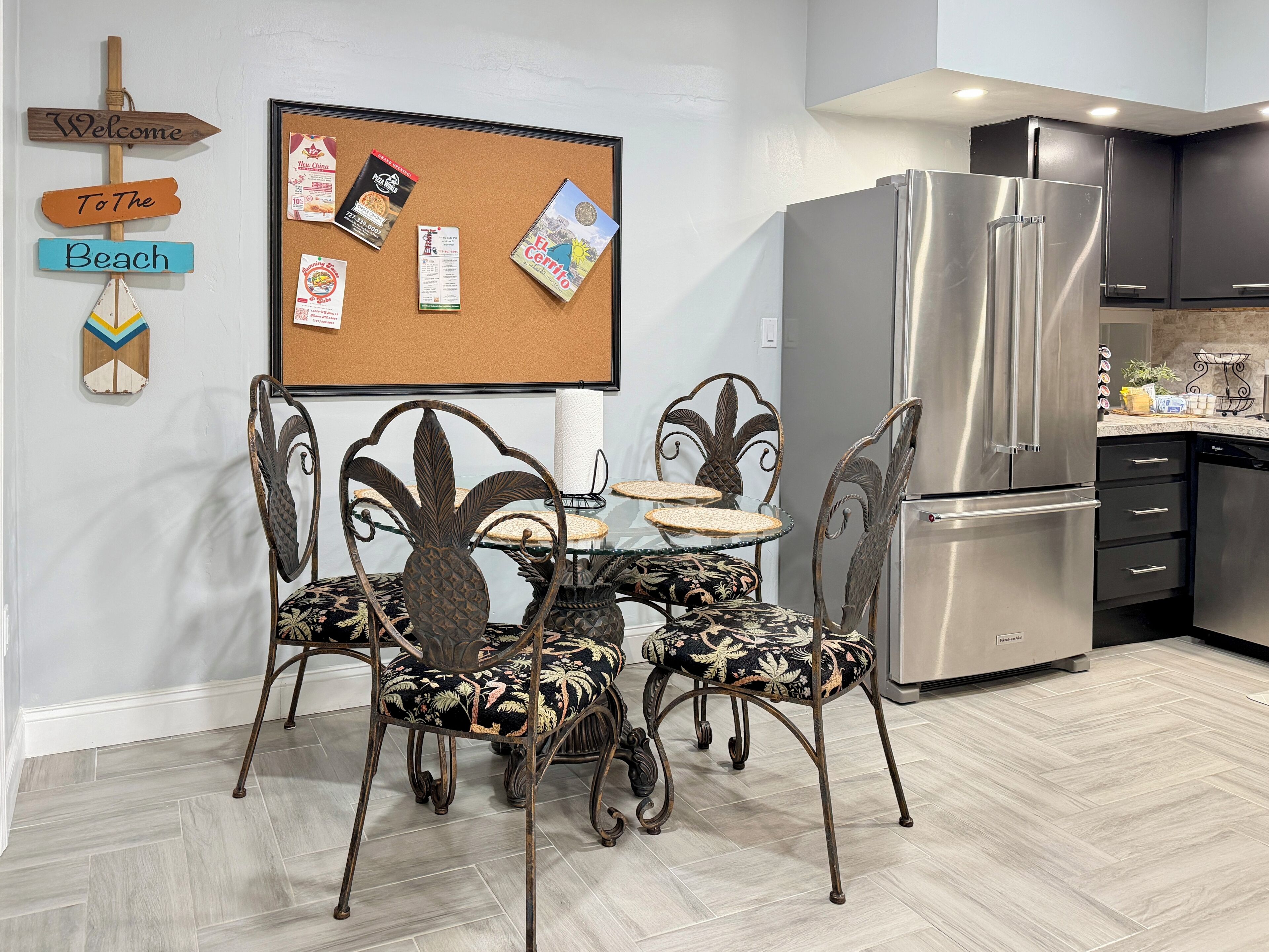 A modern kitchen with a stainless steel refrigerator, a dining table with ornate chairs, a corkboard with various notes and a welcome sign, and a herringbone-patterned floor.