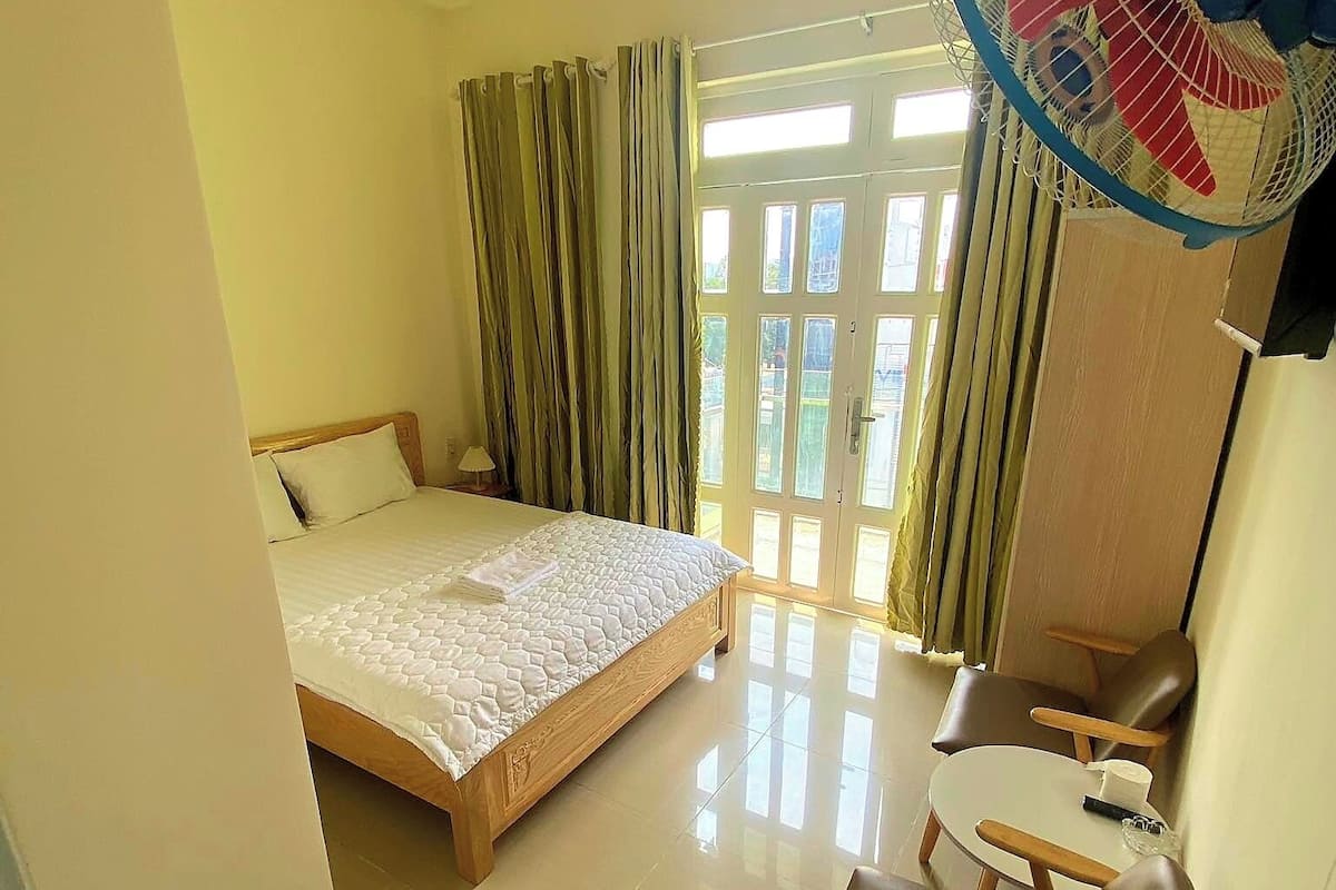 Une chambre d’hôtel avec un lit, une petite table, un canapé, un ventilateur et une paire de chaussons posés sur le sol.