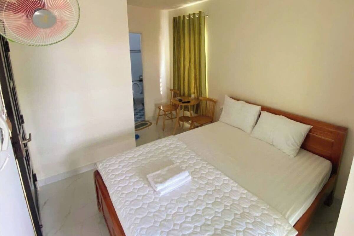 Une chambre d’hôtel avec un lit, un bureau avec des chaises, un ventilateur et une fenêtre avec des rideaux.