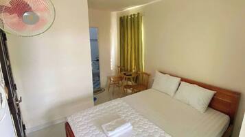 A hotel room with a bed, a desk with chairs, a fan, and a window with curtains.