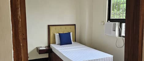 Una cama individual con cabecera de madera, una mesita de noche, una ventana con persianas y un equipo de aire acondicionado.