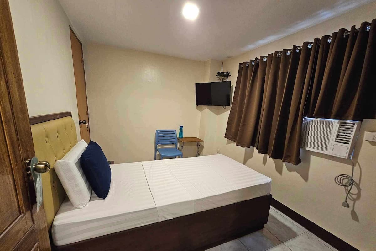 A hotel room with a bed, a television mounted on the wall, a window with curtains, and an air conditioning unit.