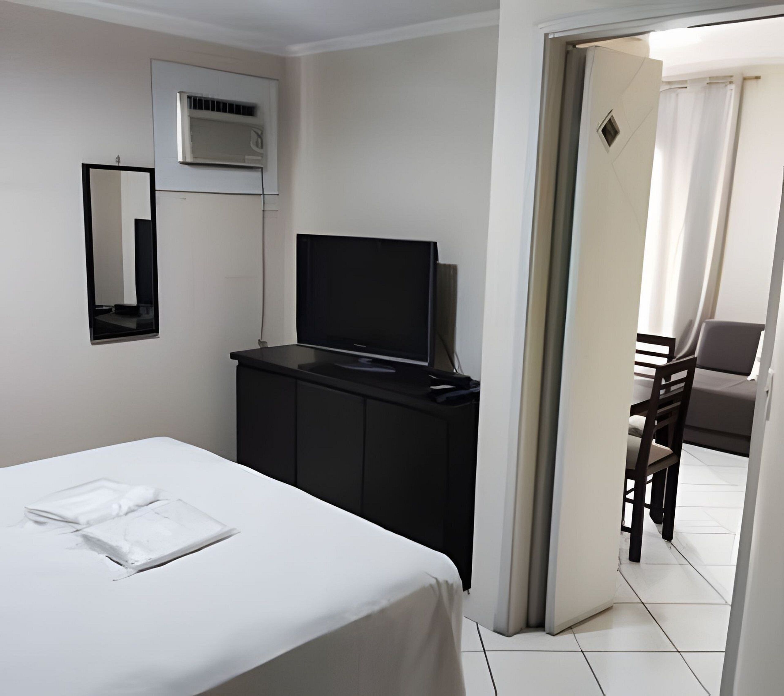 A hotel room with a bed, a TV on a black cabinet, a mirror on the wall, and a chair in the corner.
