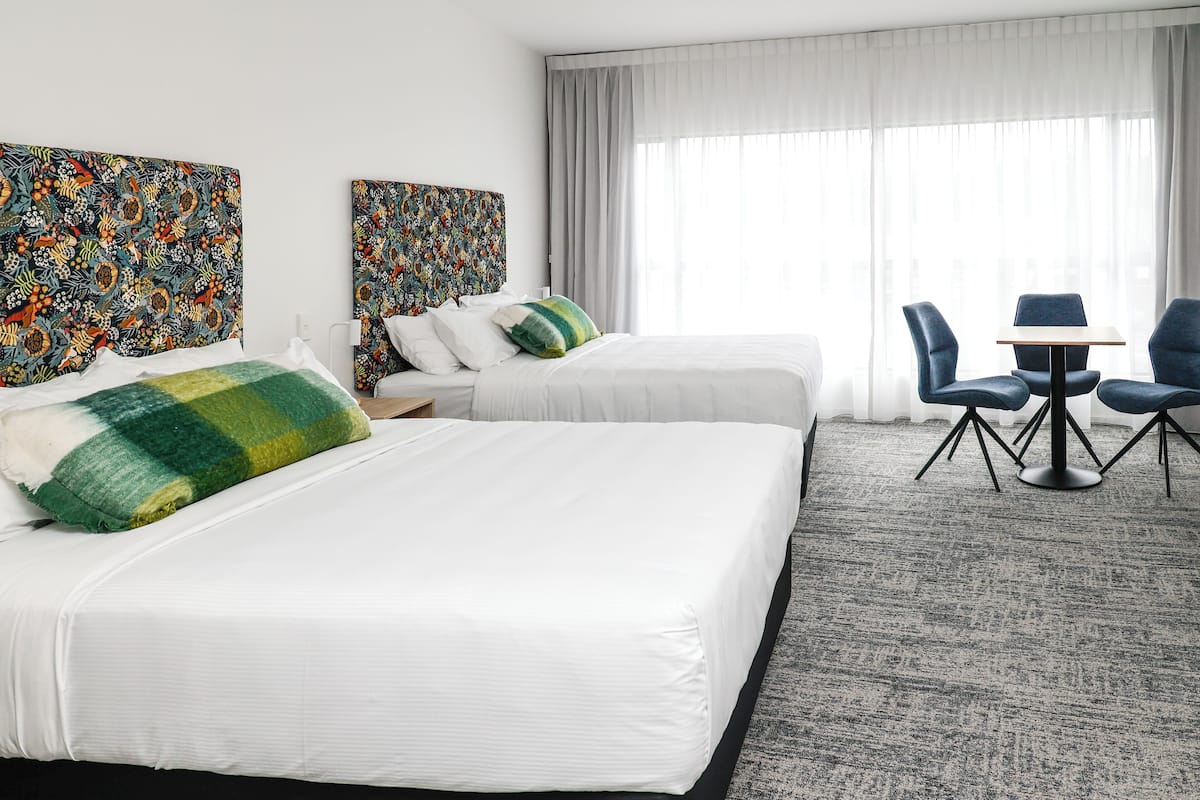 Habitación de hotel con dos camas, una mesita pequeña con sillas y ventanas amplias con cortinas traslúcidas.