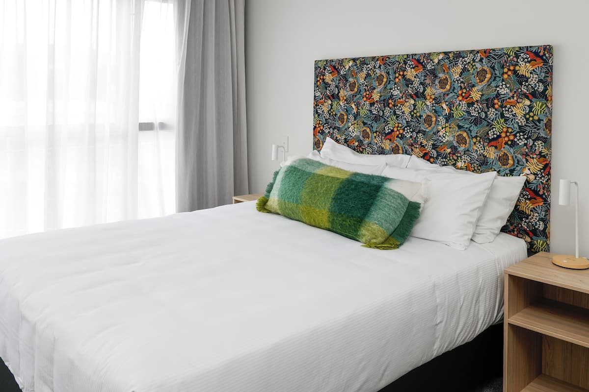 Una cama bien hecha con sábanas blancas y un cojín decorativo de colores, una mesita de noche de madera con una lámpara y un cabecero de estampado floral vibrante.
