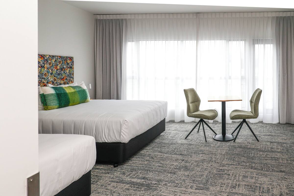 Habitación de hotel con una cama grande, dos sillas, una mesita pequeña y un ventanal con cortinas.