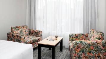 A hotel room with two floral-patterned armchairs, a wooden table with a magazine and a glass of water, and a bed with a white cover.