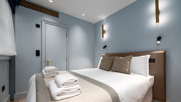 A neatly made bed with white linens and a grey accent, a wooden headboard, and two wall-mounted lights. - GuestReady - Fitzroy delight near Regent’s Park (London)