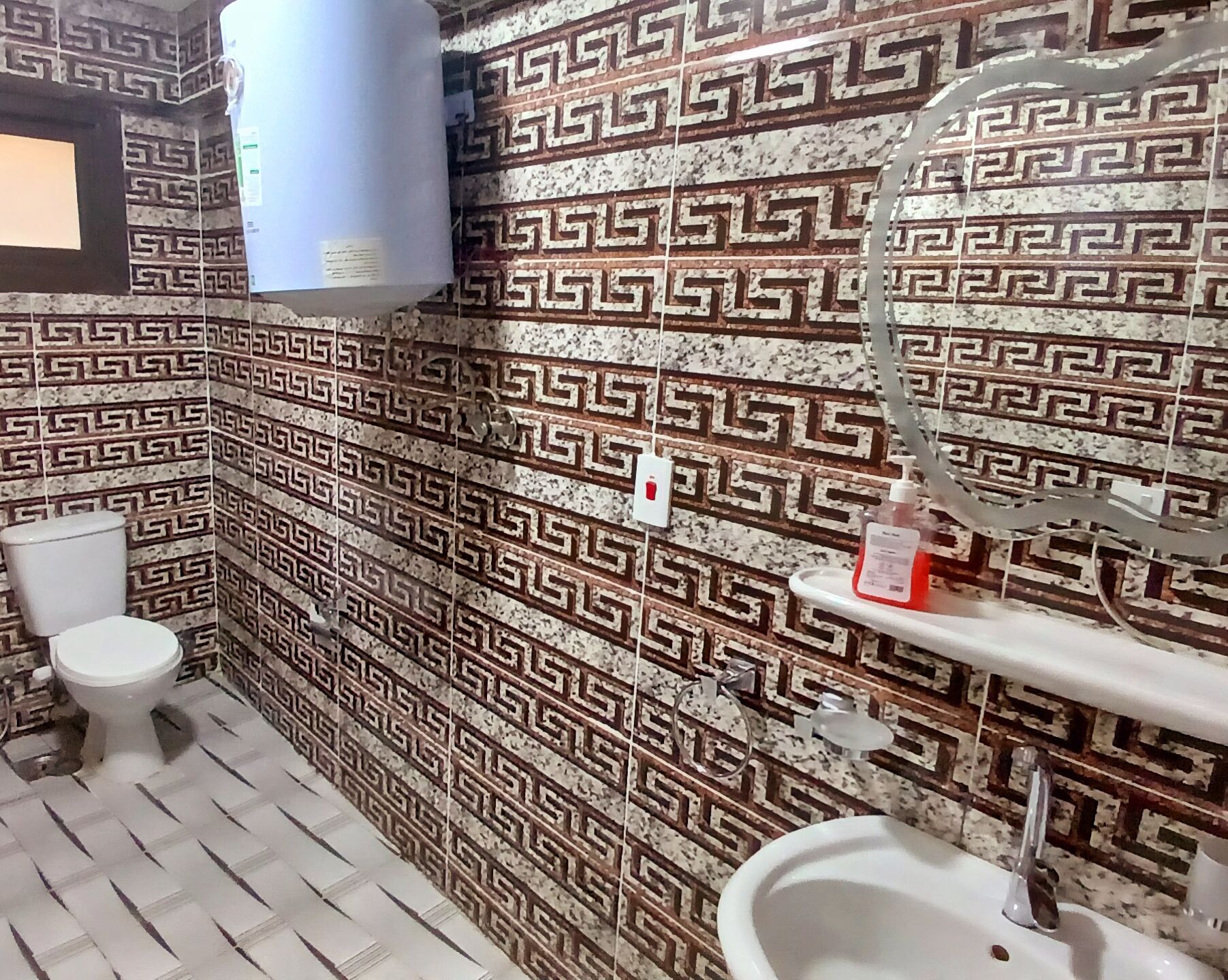 A bathroom with patterned tiles, a toilet, a bidet, a sink, a mirror, and a soap dispenser.