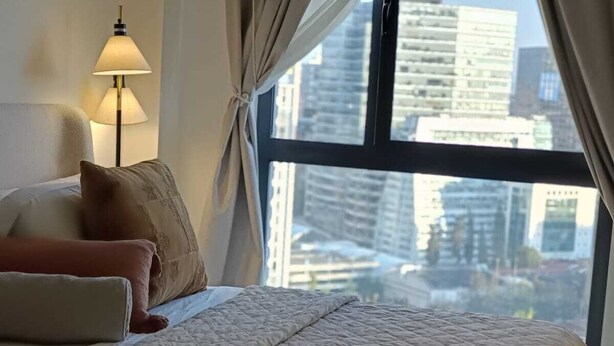 A hotel room with a bed, a lamp, and a view of tall buildings through the window. - Luxury suites polanco  (Ciudad de México)