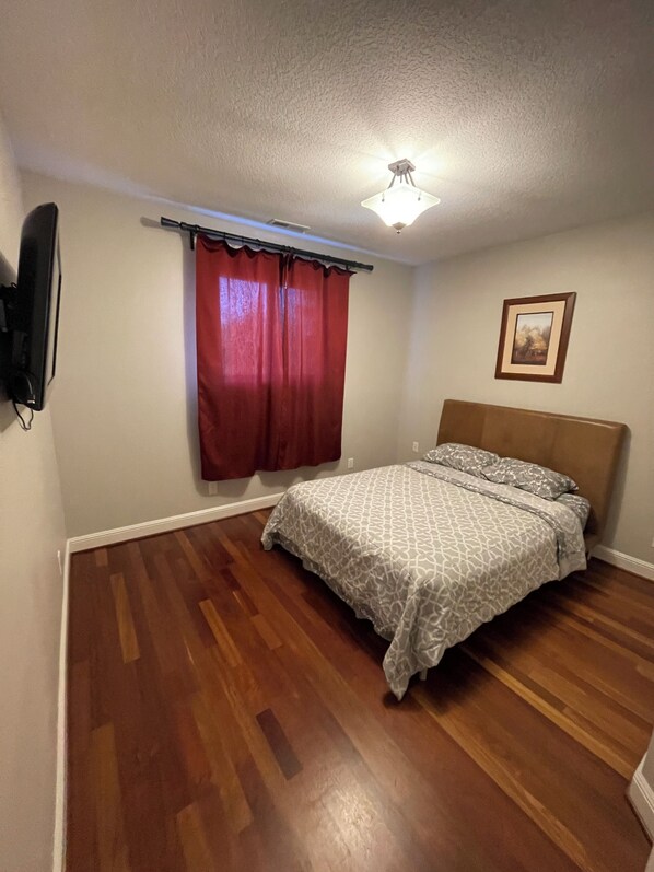 Une chambre à coucher avec un lit, un plancher en bois, un luminaire au plafond, une fenêtre avec des rideaux rouges et un tableau encadré au mur.