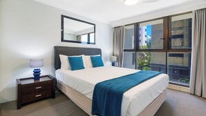 A hotel room with a large bed, a nightstand, a mirror, and a window with curtains. - Beachside 1BR Ocean View Apartment with Parking (Broadbeach)