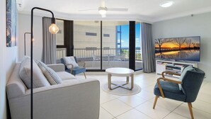 A modern living room with a sofa, chairs, a round table, and a TV. - Beachside 1BR Ocean View Apartment with Parking (Broadbeach)