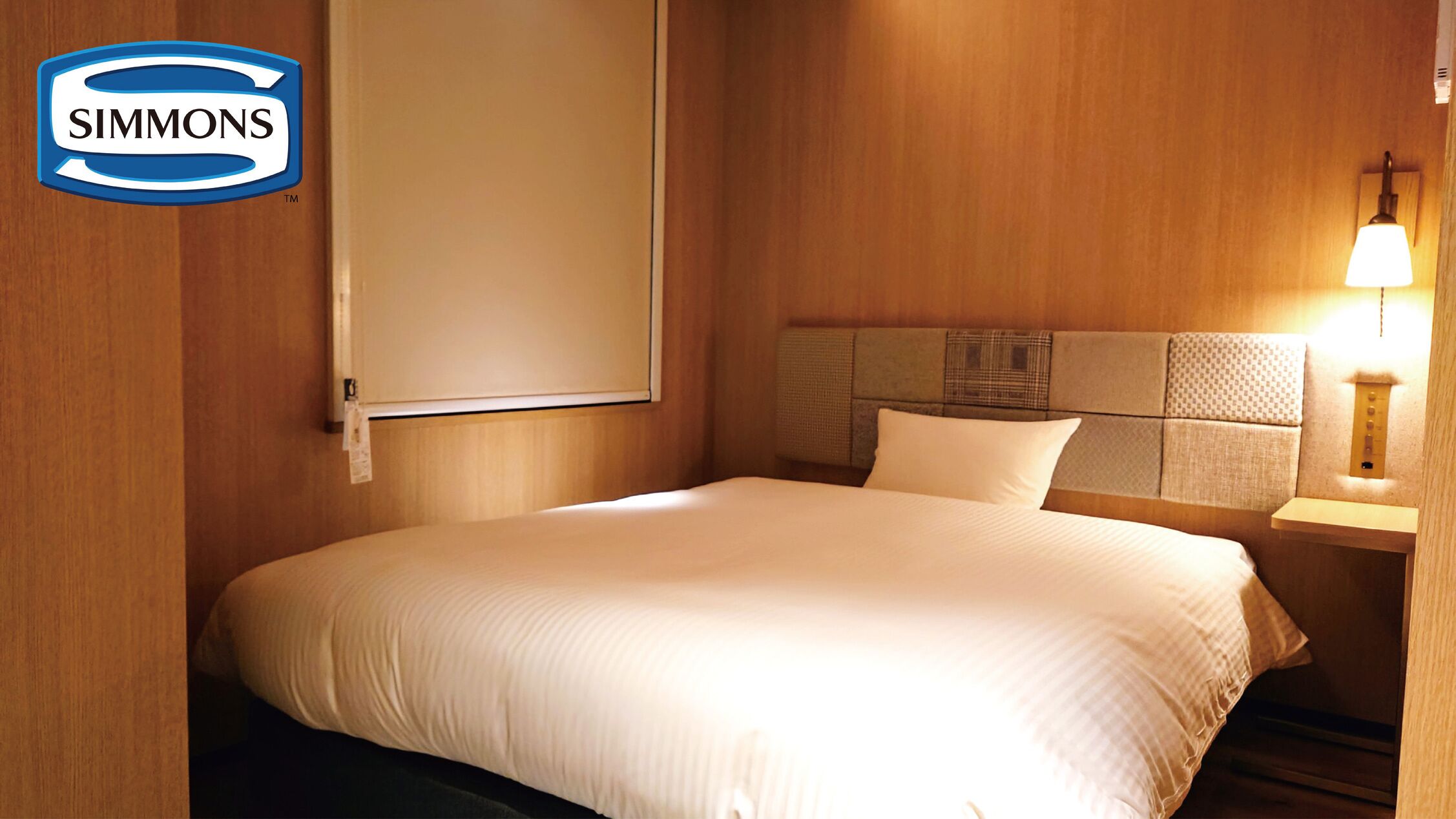 Une chambre d’hôtel de la chaîne Simmons, comprenant un lit, une table de chevet, une lampe et une tête de lit ornée d’un panneau texturé.