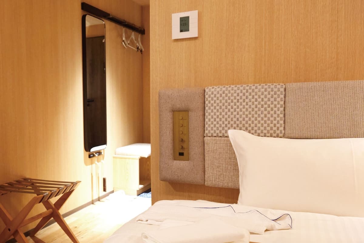 Ett hotellrum med trägolv, en säng med vita sängkläder, en trästol, ett litet bord och en väggmonterad kontrollpanel.