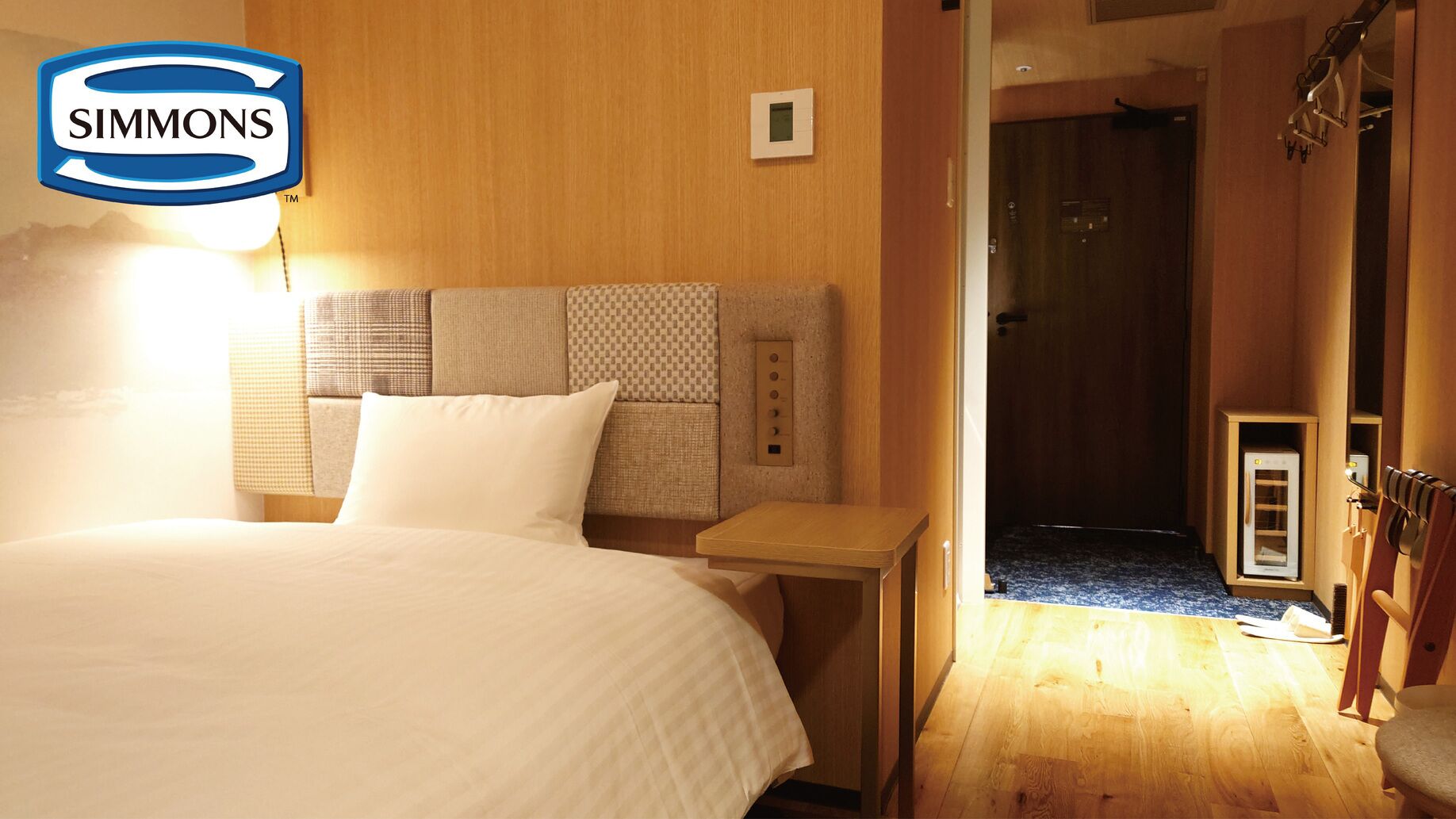 Une chambre d’hôtel comprenant un lit, une table de chevet, un panneau de commande fixé au mur et une porte donnant accès à un espace douche.