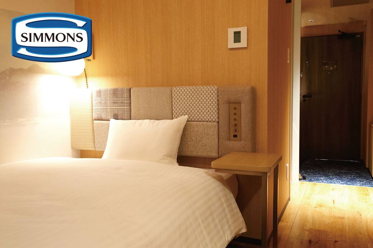 Une chambre d’hôtel comprenant un lit, une table de chevet, un panneau de commande fixé au mur et une porte donnant accès à un espace douche.