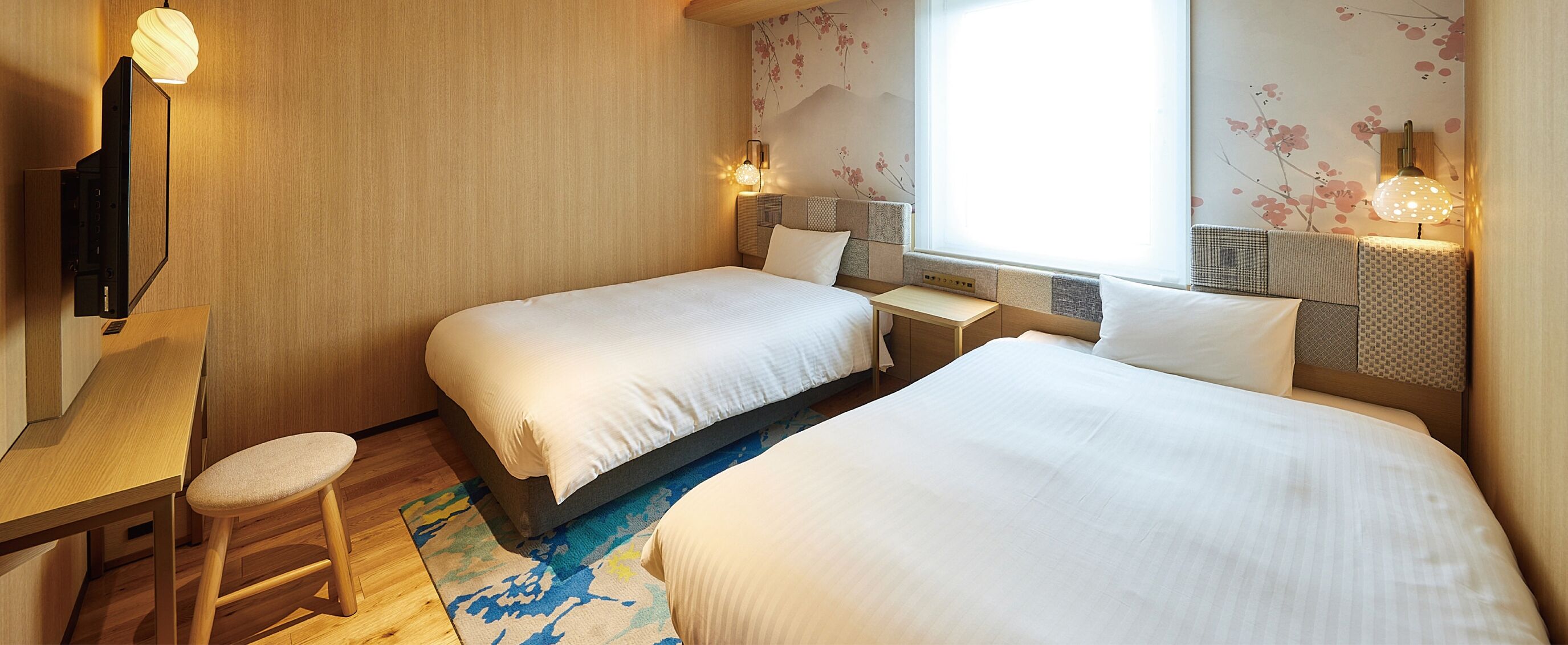 A hotel room with two beds, a desk, and a chair. There are wall-mounted lamps and a large window with a view of cherry blossoms.