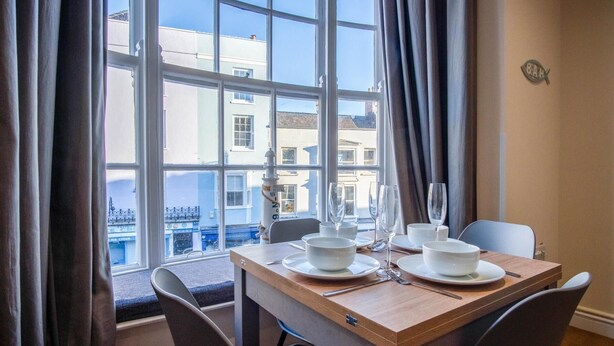 Un comedor con una mesa lista para dos, vistas de edificios a través de ventanas amplias, y un cartel que dice "NEIGHBORHOOD BEACH".