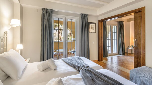 A hotel room with two beds, a balcony with curtains, a wooden door, and a view of the outside.