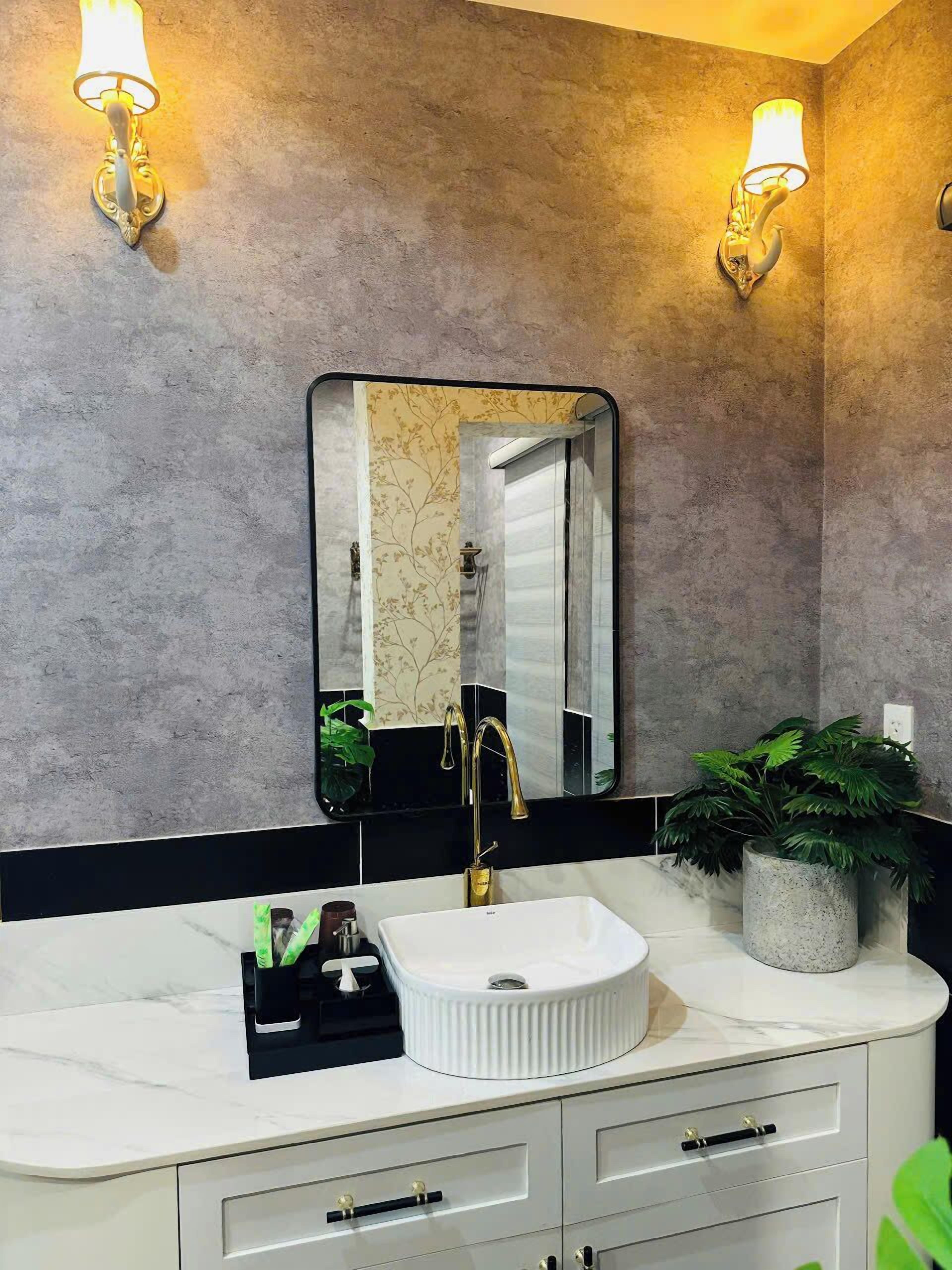 A bathroom with a white sink, gold faucet, and a large mirror. There are two wall-mounted lights and a potted plant on the counter.