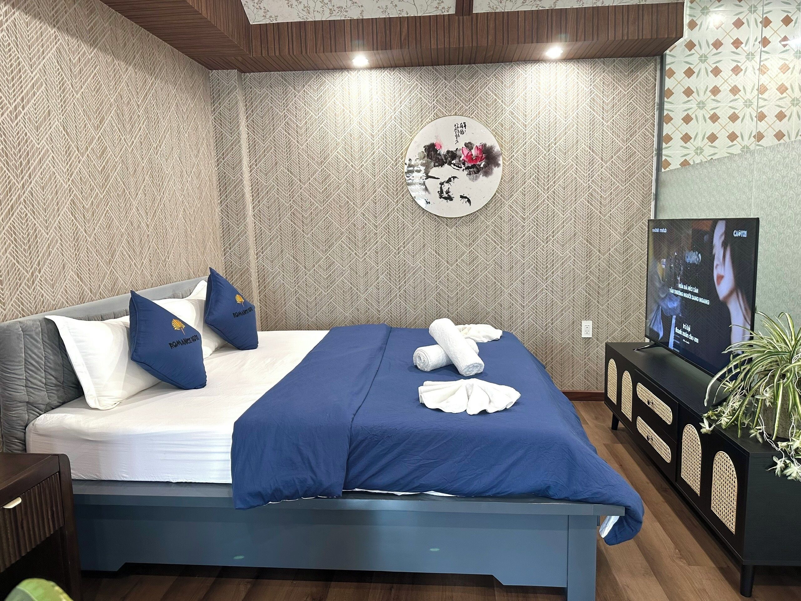 Een net opgemaakt bed met een blauw sprei en kussens, een muur met een geometrisch patroon, een decoratief rond object en een televisie op een standaard.