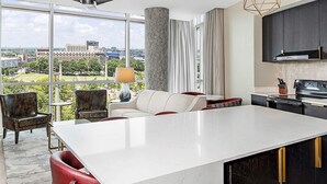Una habitación de hotel moderna con un ventanal grande, una isla central, una zona de cocina y un área de estar con vistas a la ciudad.