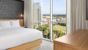 Una habitación de hotel con una cama grande, un cabecero de madera, una mesita de noche, un escritorio y vistas a la ciudad a través de amplios ventanales.