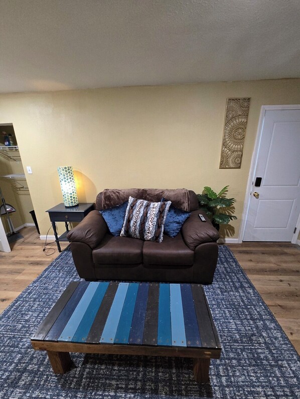 A living room with a brown sofa, a striped coffee table, a patterned rug, a small side table with a lamp, and a decorative wall piece.