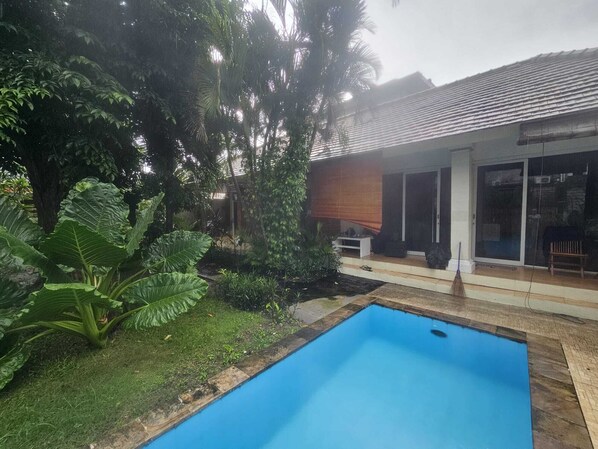 A swimming pool with a wooden deck, surrounded by lush greenery and a house with a tiled roof. - Two Brothers Guesthouse (Legian)