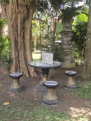 A garden setting with a table and stools, a laptop on the table, and a large tree in the background. - Two Brothers Guesthouse (Legian)