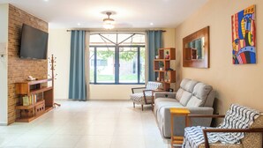 A living room with a TV mounted on a brick wall, a sofa, a chair, a wooden bookshelf, and a large window with curtains. - Apt with Pool & jungle in Puerto Morelos (Puerto Morelos)