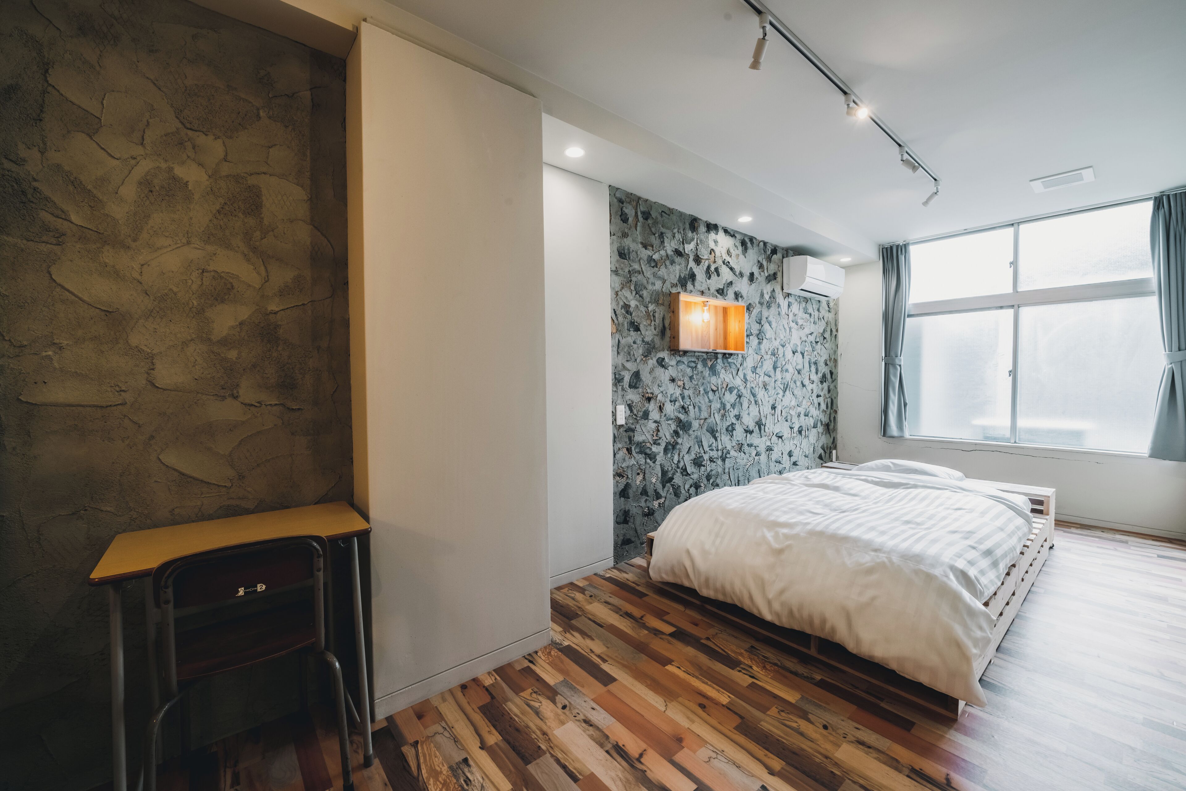 Un dormitorio con pared de piedra, un escritorio de madera, una cama con colcha blanca y una ventana con cortinas.