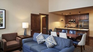 Un salon avec un canapé bleu, deux coussins à motifs, un fauteuil marron, une petite table avec une lampe, et un coin repas avec une table et des chaises en bois.