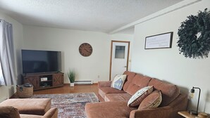 Una sala de estar con un sofá marrón, dos sillones, un televisor de pantalla plana grande, un aparador de madera, un reloj de pared decorativo, una cita inspiradora enmarcada y una pequeña planta en maceta.